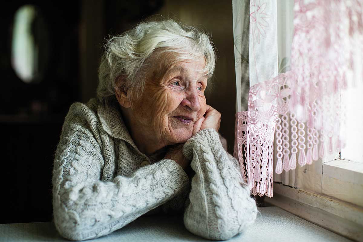 elderly woman looking outside of window with pink curtain and white sweater
