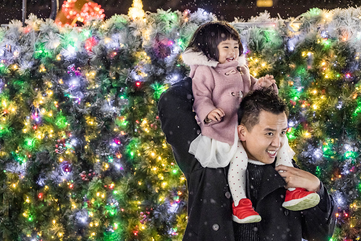 father with daughter on shoulder in front of bushes with holiday lights