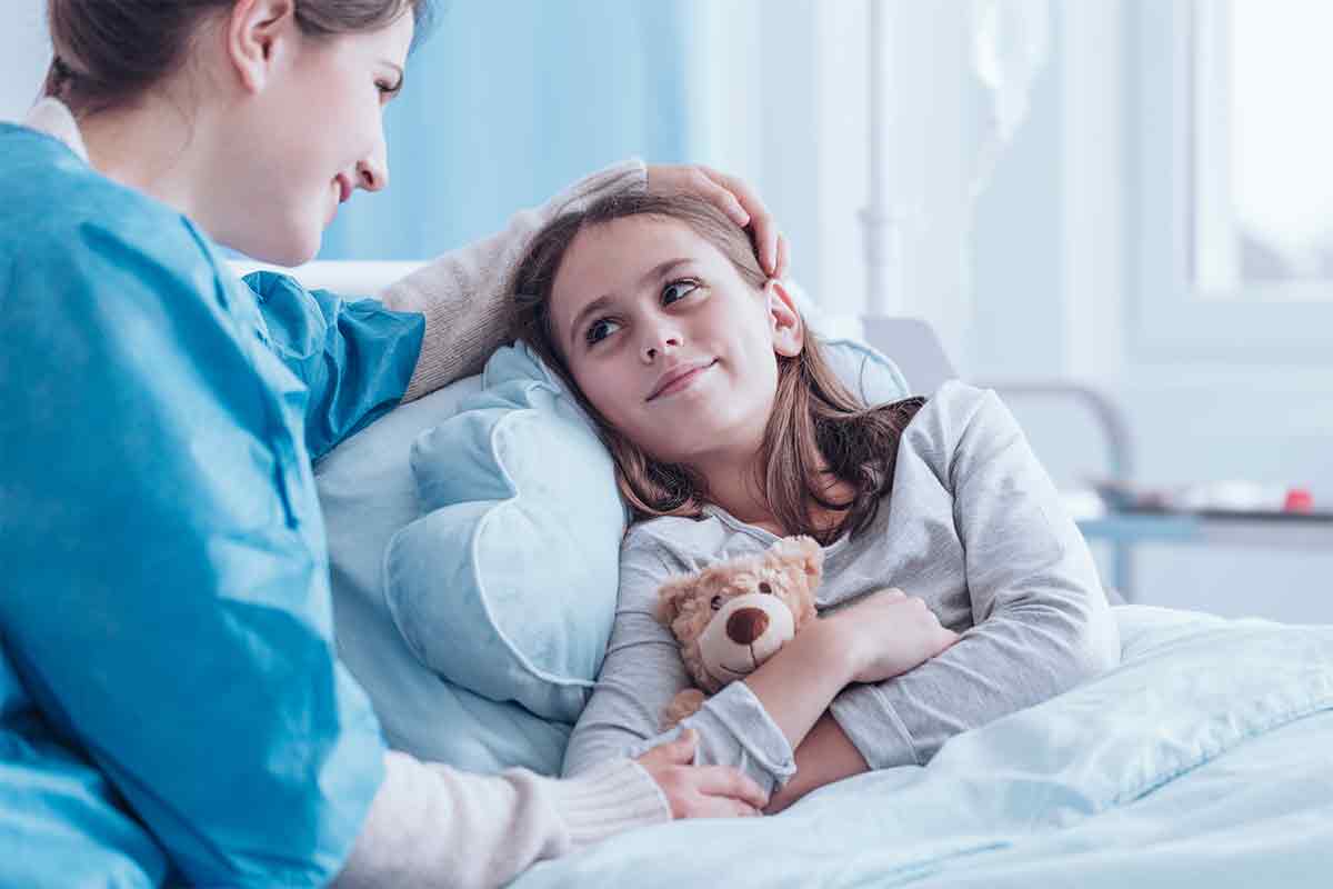 child holding a teddy bear in hospital bed as nurse comforts her