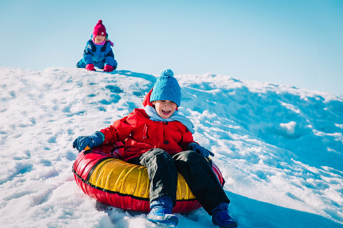 kid sledding down a snowy hill with blue hat and red tube