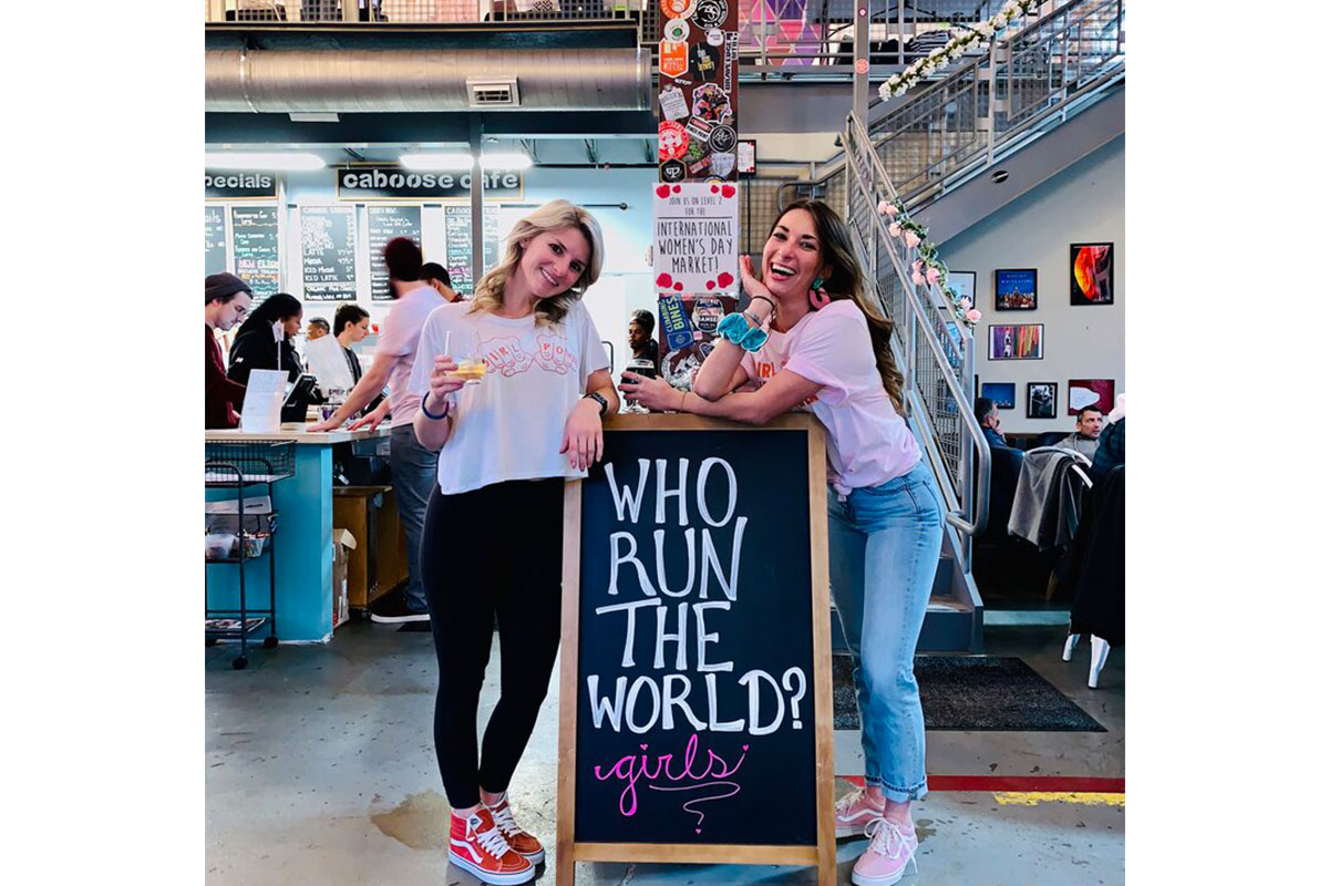two women by a sign that says who run the world?