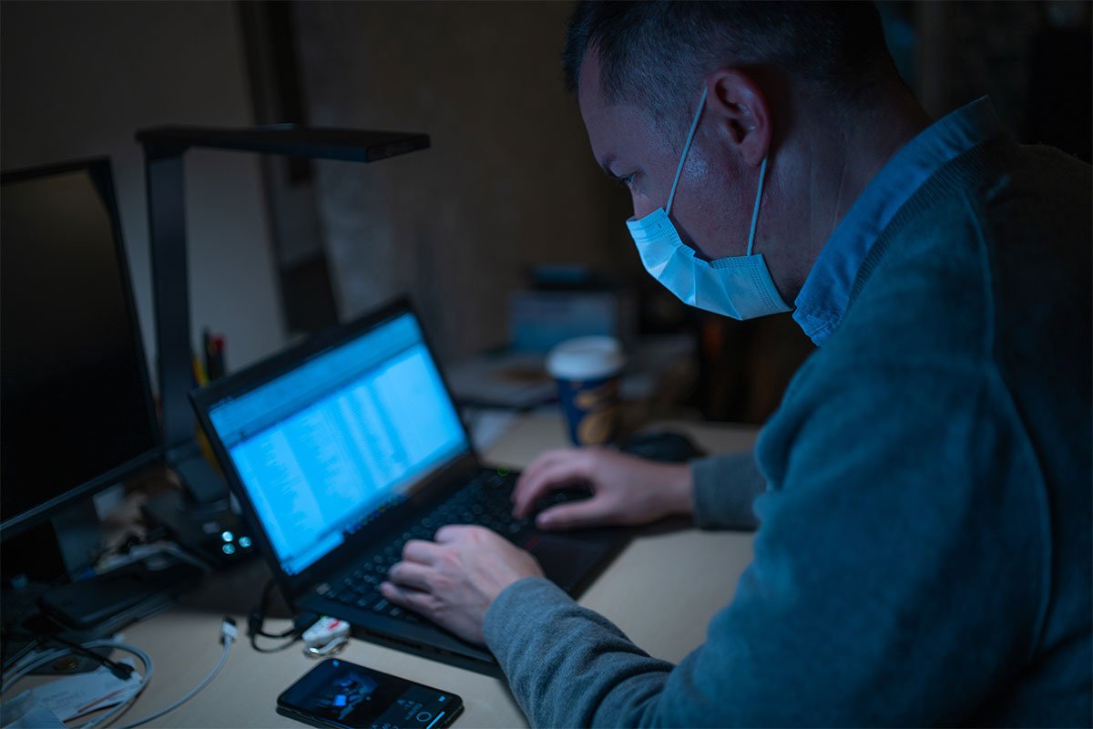 man wearing blue medical mask looking at blue laptop screen in dark room