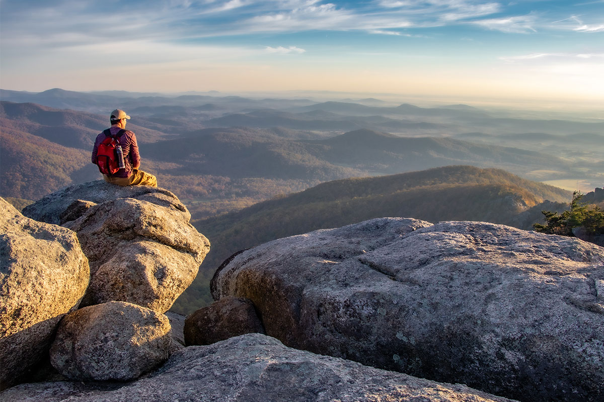 man sitting on rock and looking out at old rag hiking with mountains and sunset and blue sky