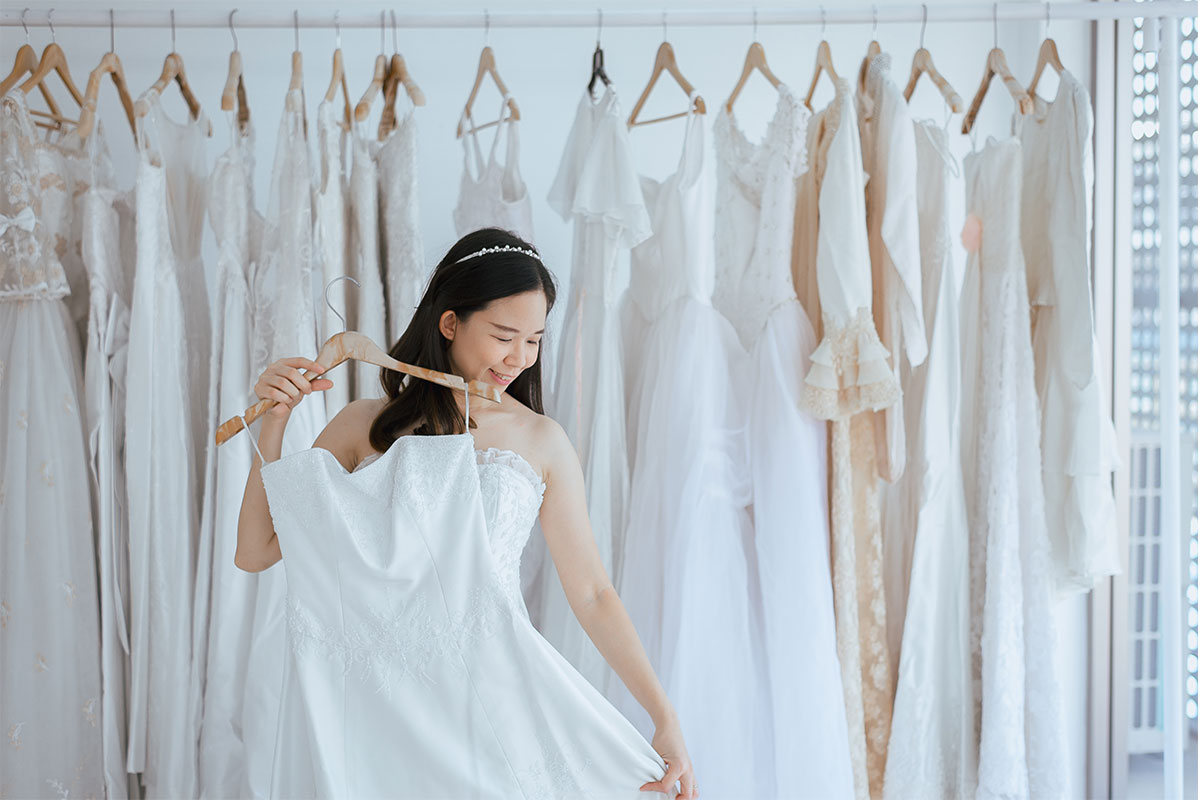 woman holding white wedding dress near window with rack of white wedding dresses behind her