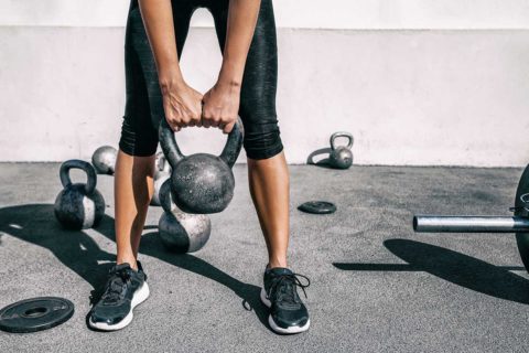 woman lifting kettlebell with other weights outside with black pants and black shoes