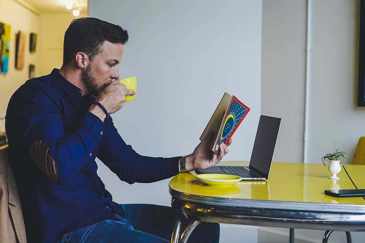 man sitting in front of laptop with yellow table holding book and yellow coffee mug