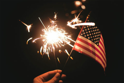 hand holding american flag with firework going off in the sky in the background