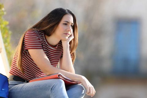 girl with brown hair sitting in maroon striped shirt with folders and jeans looking sad