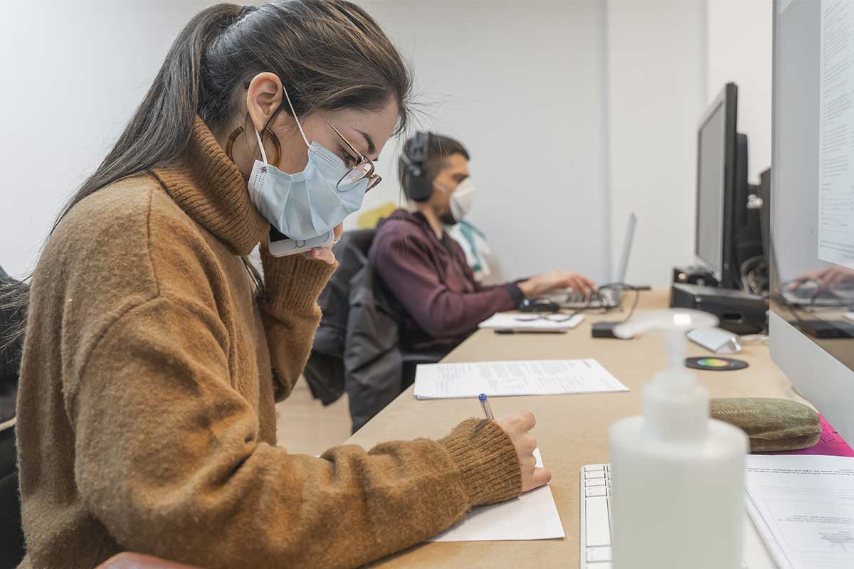 college student in brown sweater wearing mask working in front of computer