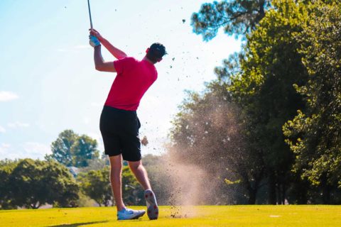 man in pink shirt golfing in front of trees and blue sky