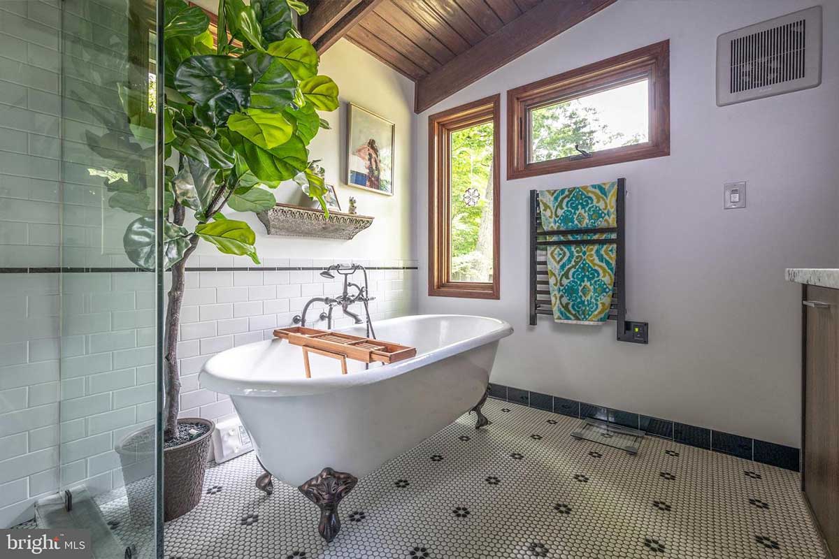A white clawfoot bathtub sits in the corner of a black and white tiled bathroom with a green tree looming over. 