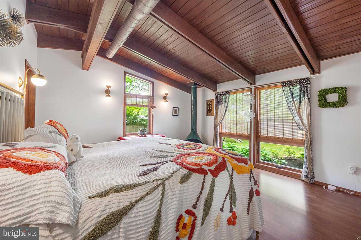 Inside a white bedroom, a white and floral bed faces two floor to ceiling windows and a fireplace rests in the corner.