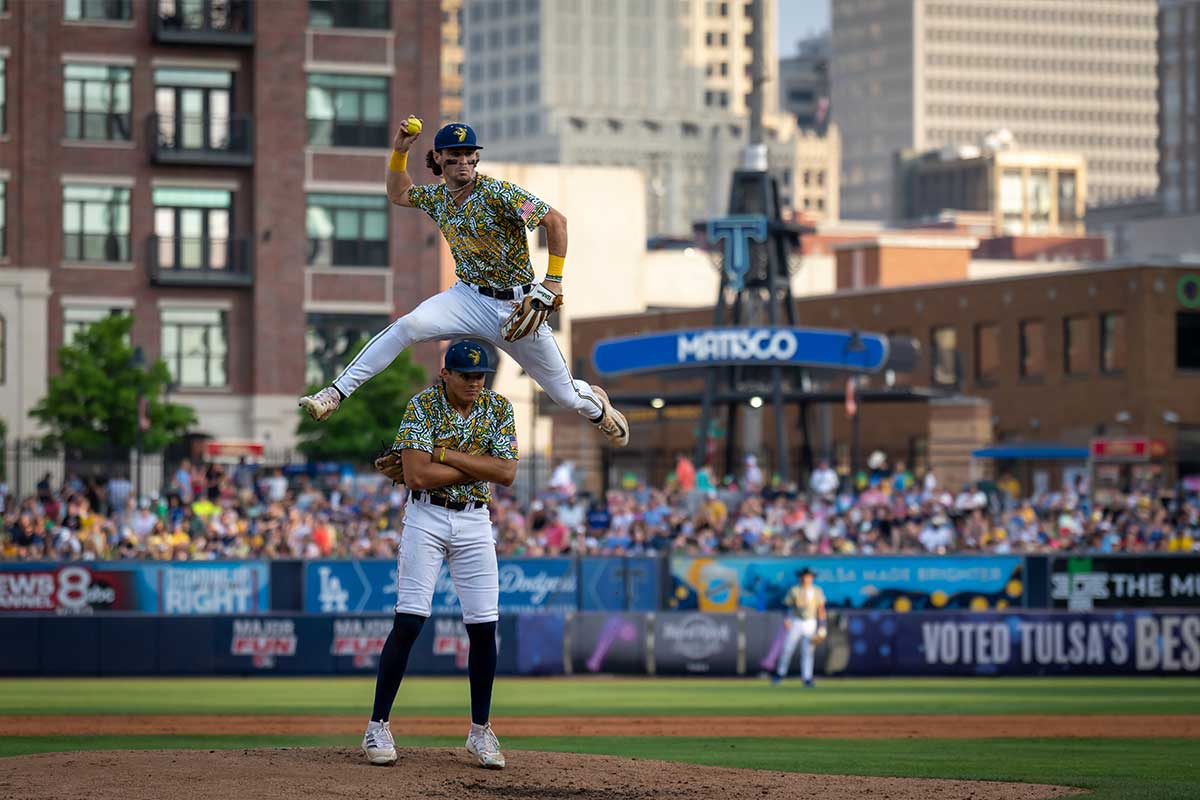 baseball player jumping over pitcher while throwing ball
