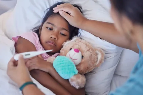 sick kid cuddled with teddy bear while mom takes temperature