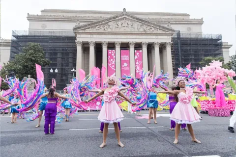 young dancers and performers in pink, purple and bright blue costumes spin colorful ribbons in front of the National Archices