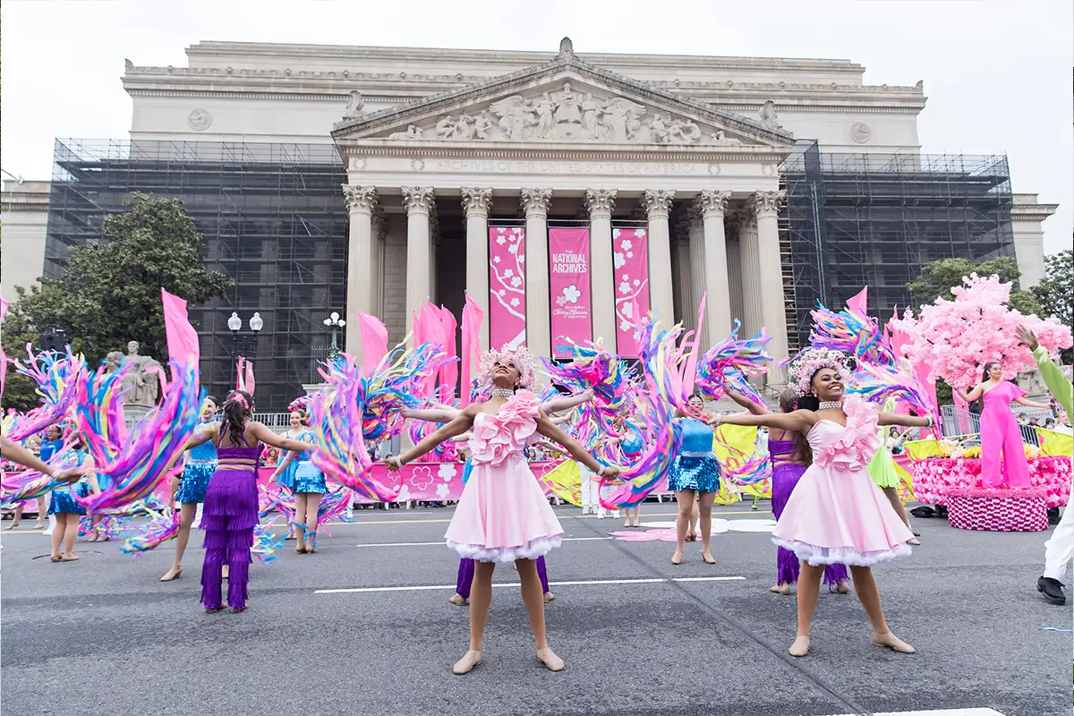 young dancers and performers in pink, purple and bright blue costumes spin colorful ribbons in front of the National Archices