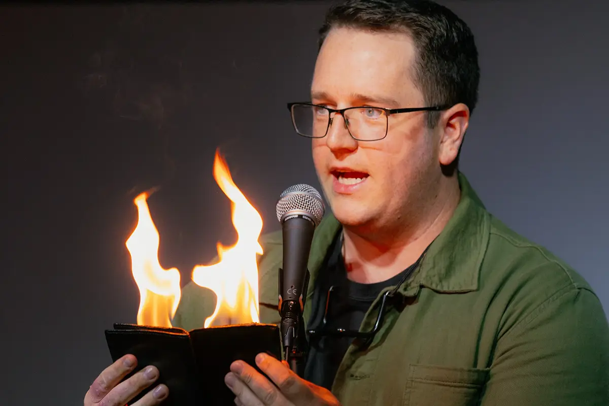 Charlie Ross stands on stage in front of a microphone holding a wallet with flames coming out of it