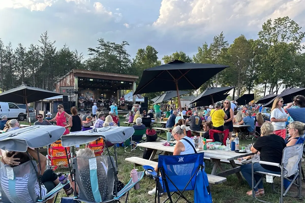 people sit on chairs and at picnic tables on the lawn while a band plays on a stage in the background