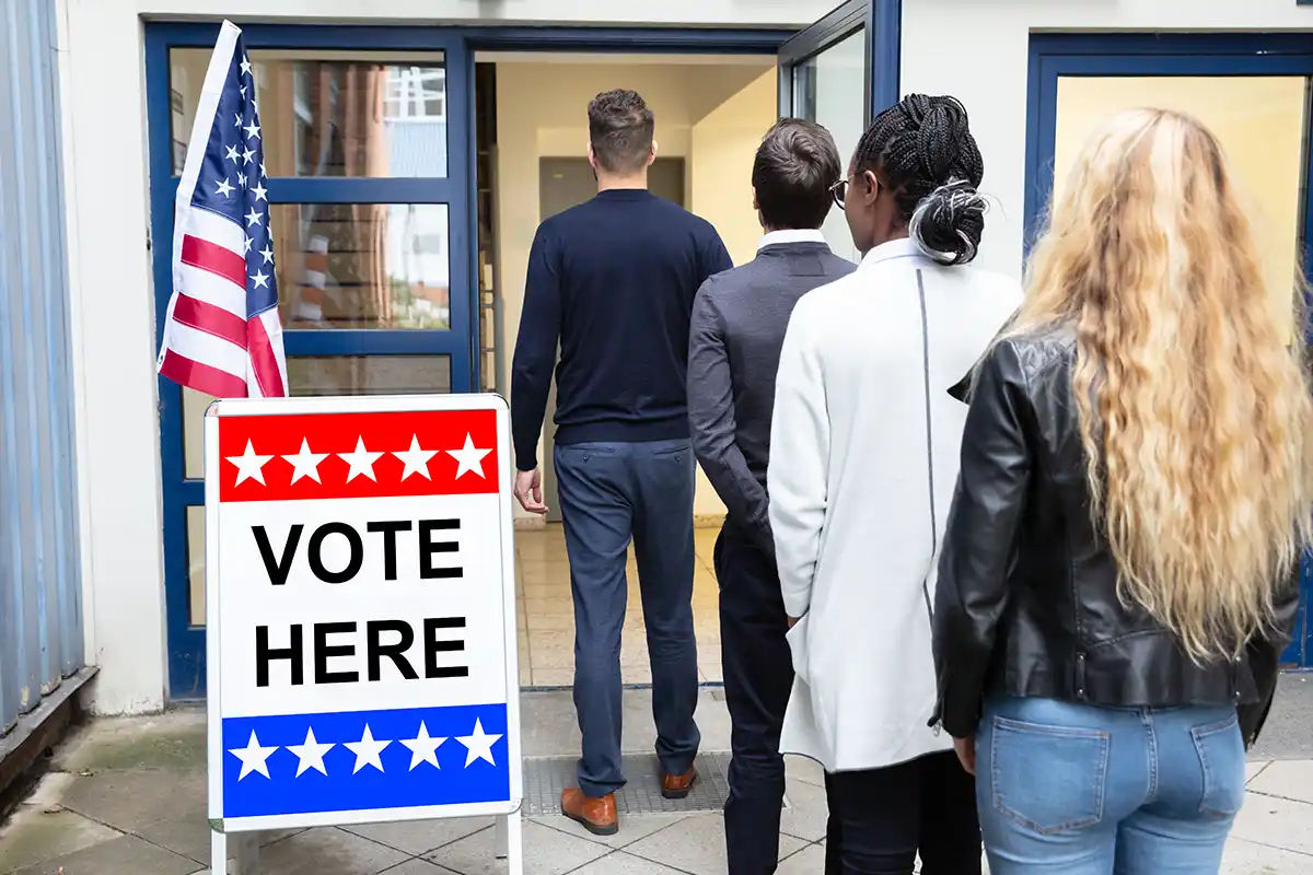 four people stand in line next to a Vote Here sign