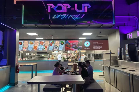 people sit at tables eating food with a neon pop-up district sign in the background