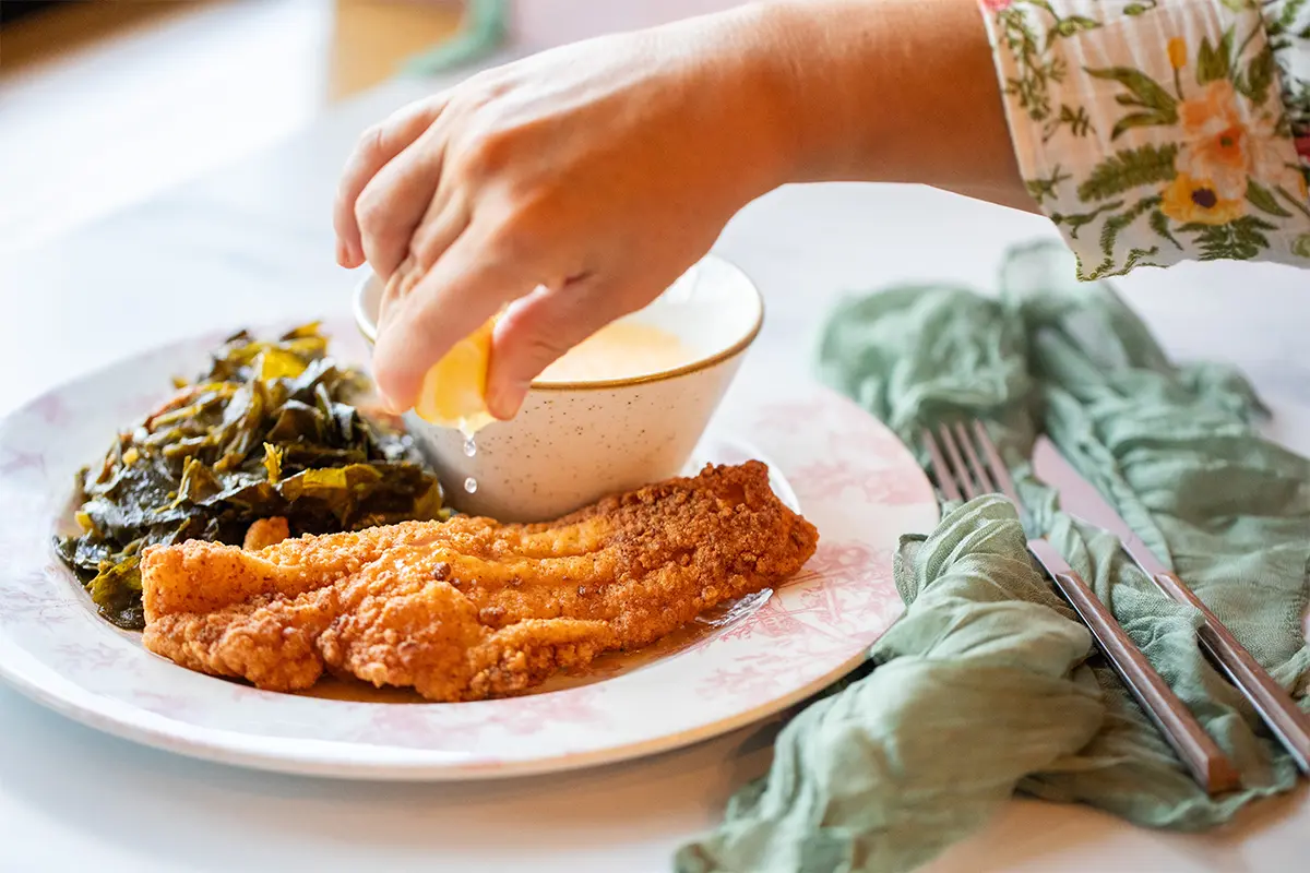 a hand squeezes a lemon on a plate of collard greens and friend chicken