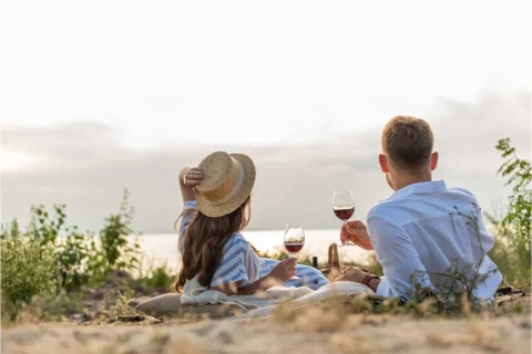a couple sit in front of a body of water sipping wine