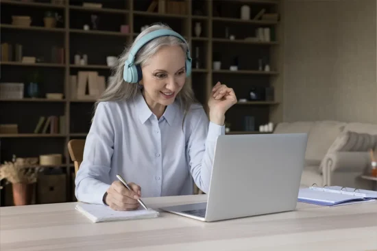 a woman wears headphones and takes notes while looking at a laptop