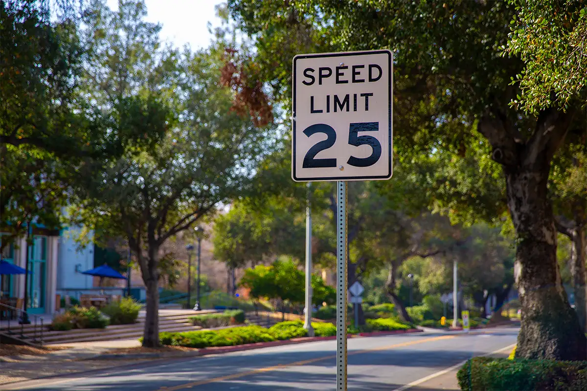 a 25 mph speed limit sign along a road with trees and buildings in the background