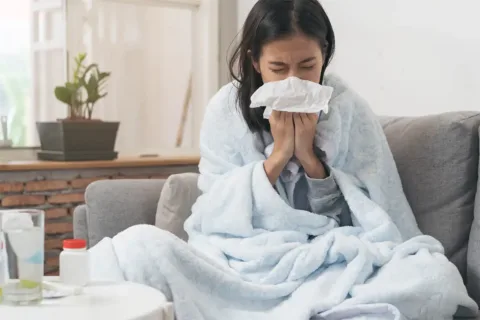 a woman sits on a couch blowing her nose with water and medicine on a nearby table
