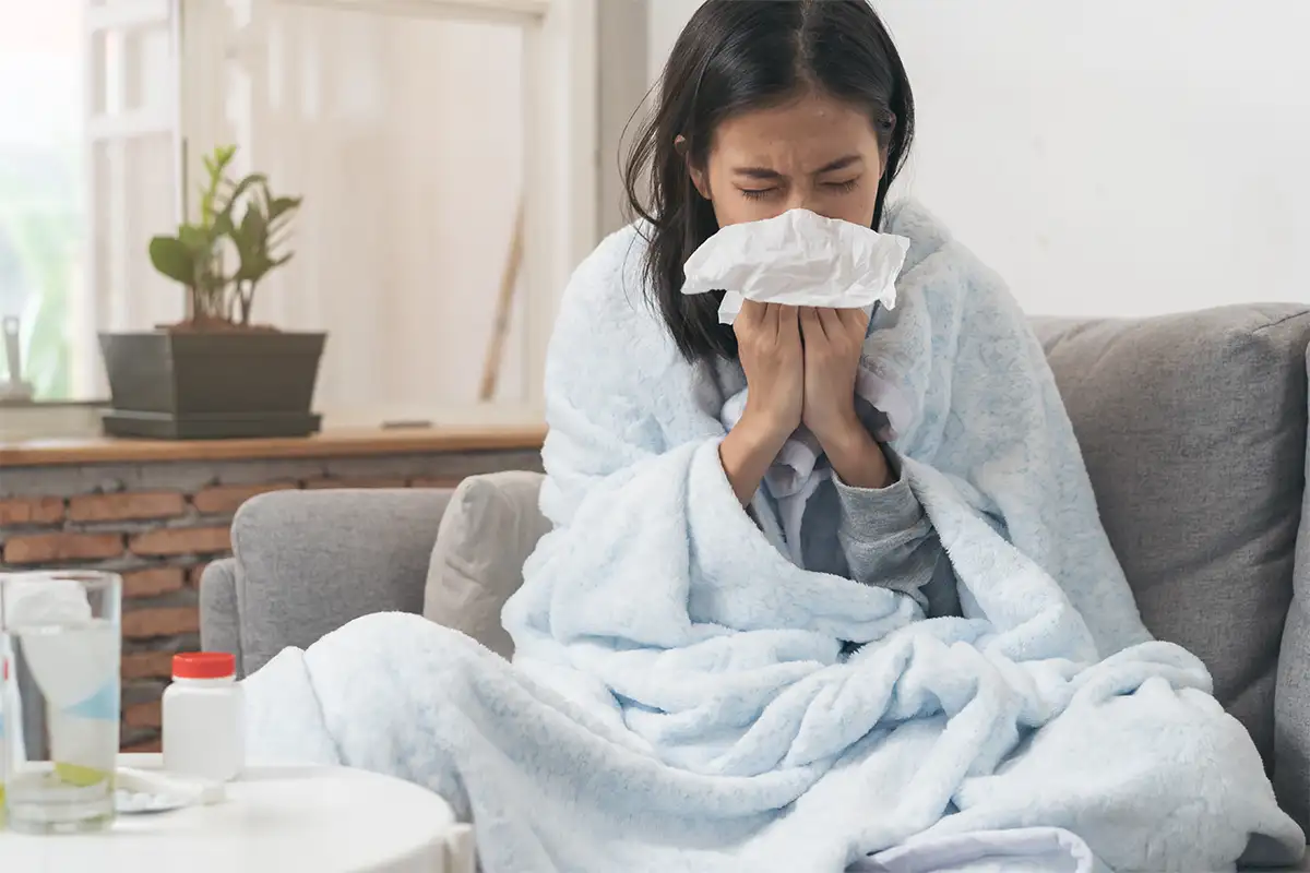 a woman sits on a couch blowing her nose with water and medicine on a nearby table