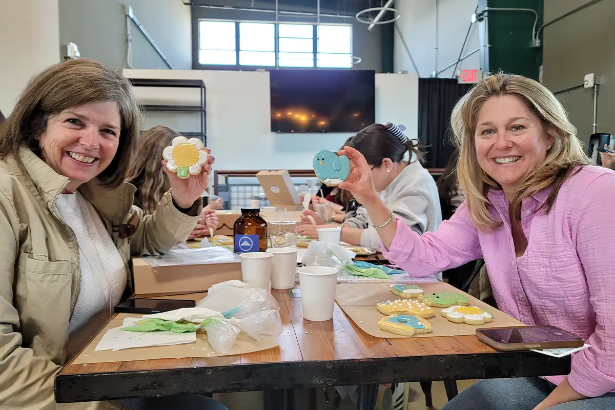 two women hold decorated cookies at a decorating class while smiling at the camera