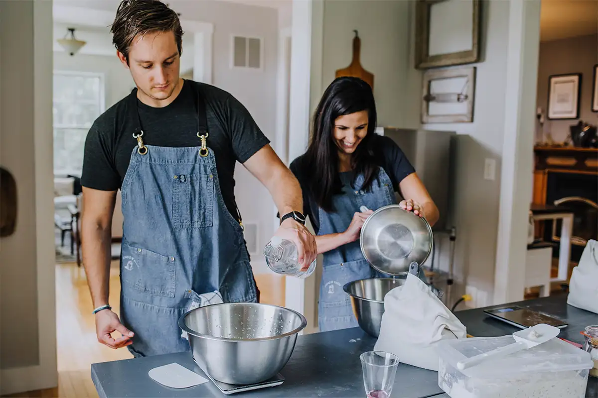 a man and woman in denim aprons standing in a kitchen mixing ingredients in a bowl