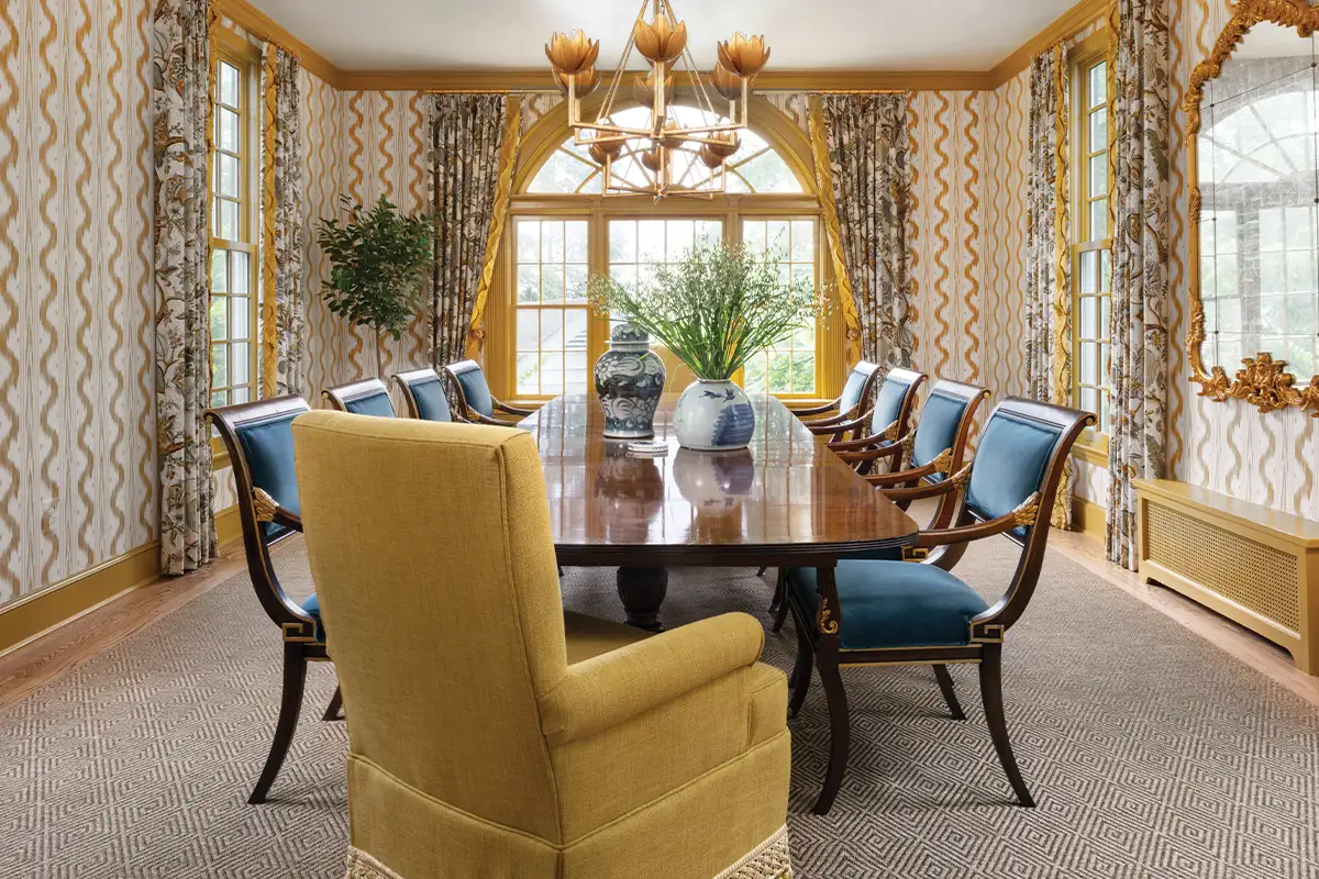 The dining room features Pierre Frey patterned wallpaper and custom drapery, edged in a Scalamandre ruffle trim. A pair of Julie Neill chandeliers hang above the table.
