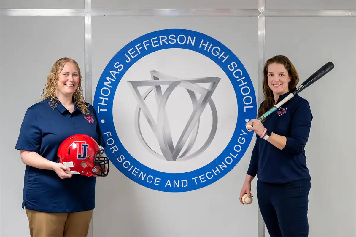 Thomas Jefferson head football coach Rachel Worsham (left) holds a football helmet and and Jennifer Hammond (right) holds a baseball bat with the TJ High School emblem between them