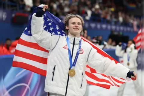 Ilia malinin skating with gold medal and flag at 2026 Olympics