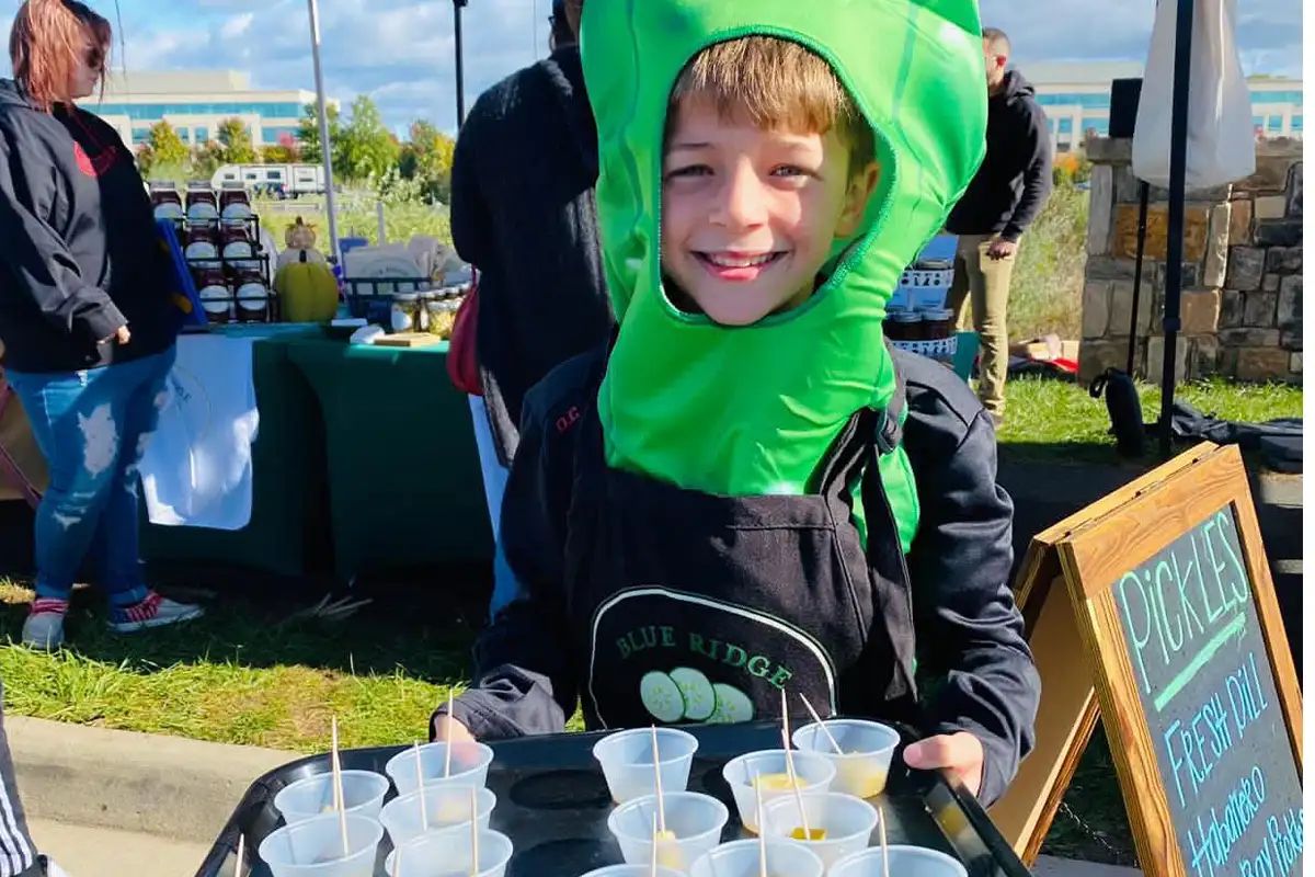 a young boy in a pickles costume holds a tray of food samples at the outdoor Pickle & International Food Festival