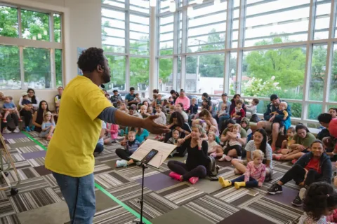 a man in a yellow shirt stands at a lectern in a room animatedly talking to parents and small children
