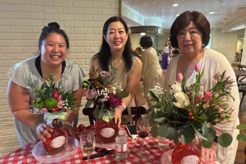 three women smile at the camera showing their floral arrangements on a table