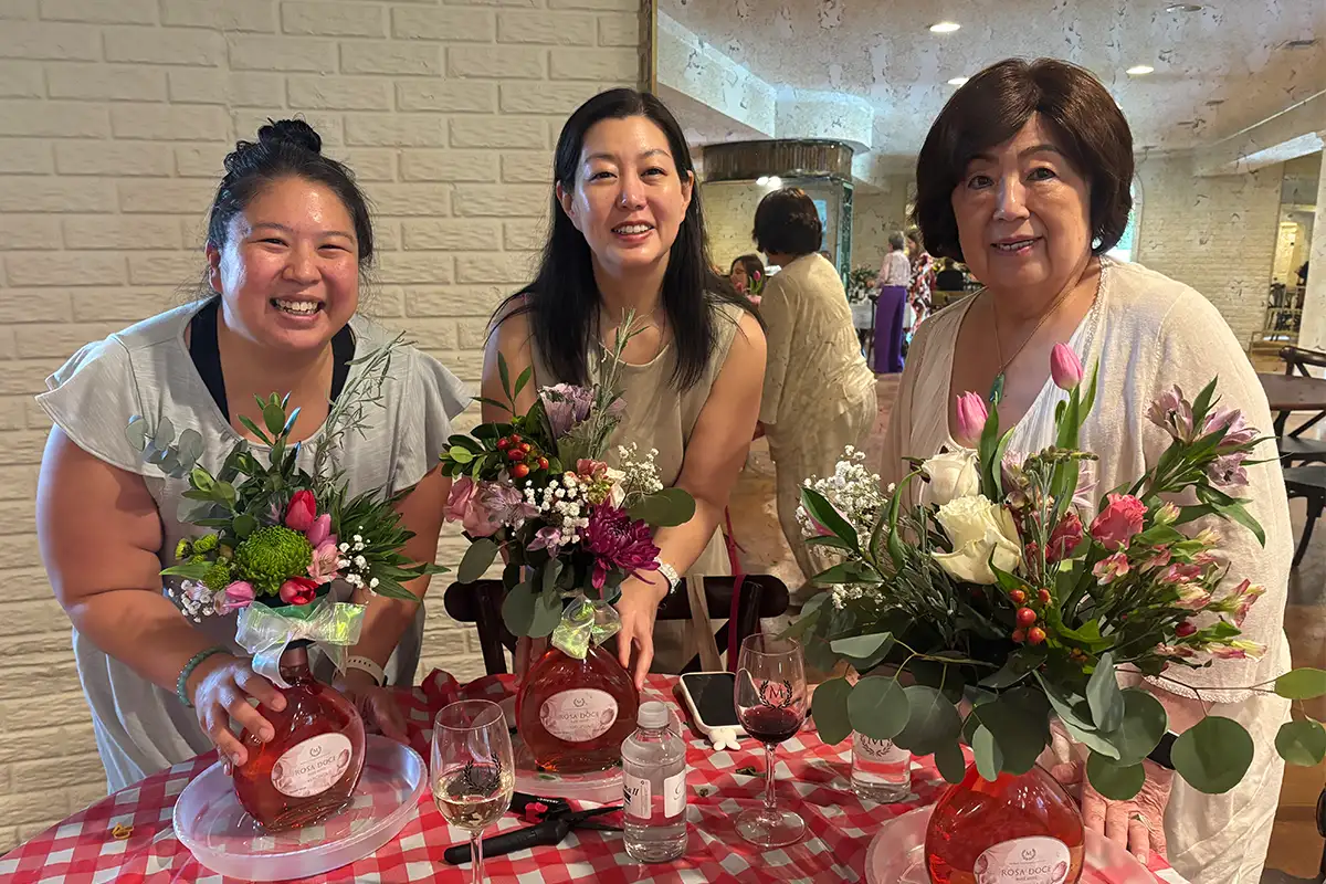 three women smile at the camera showing their floral arrangements on a table
