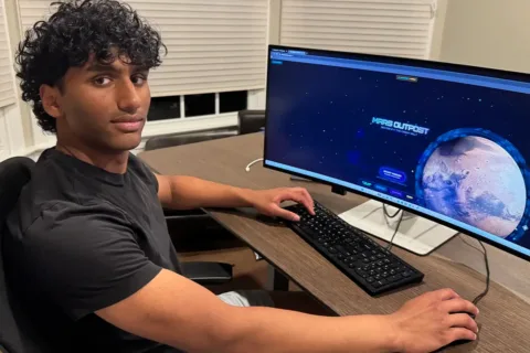 Rishab Nanduri sits in front of a computer that shows his MoneyQuest website