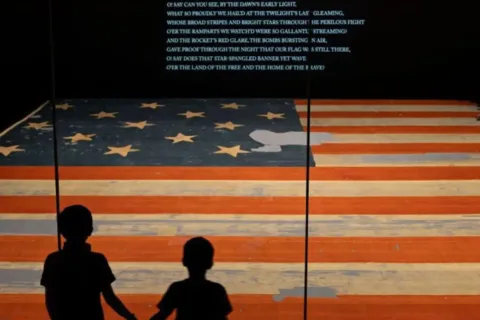 two small children stand in front of an American Flag as part of the National Museum of American History Commemorates the 250th With an Exhibition That Spans the Entire Museum