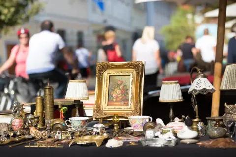 items on a able including art, metal collectables, lamps, and china at an outdoor flea market sale with people in the background