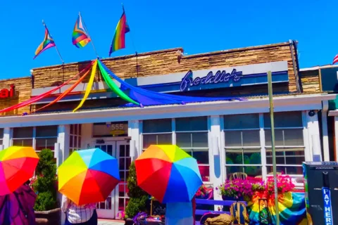 Three people standing outside of Freddie's Beach Bar with rainbow umbrellas