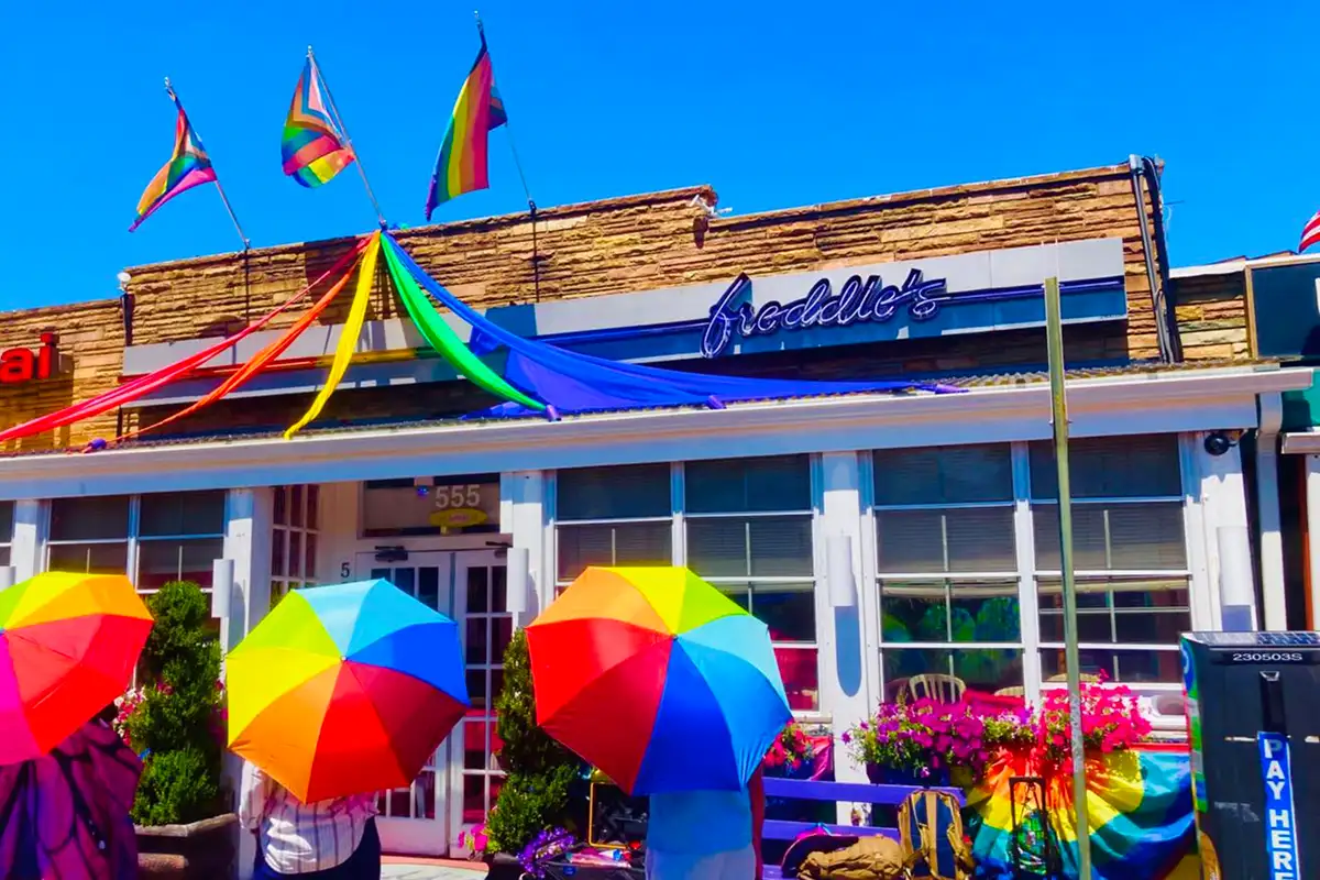 Three people standing outside of Freddie's Beach Bar with rainbow umbrellas