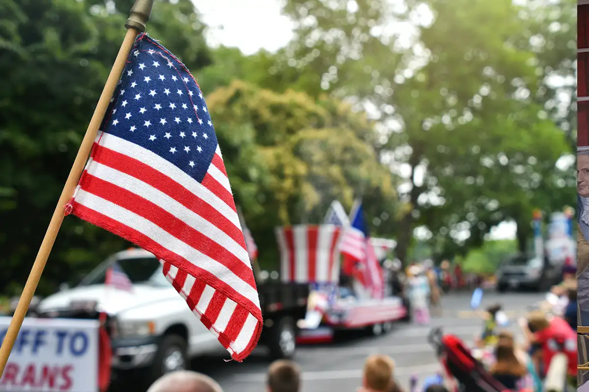 an American flag waves in front of the blurred image of a parade