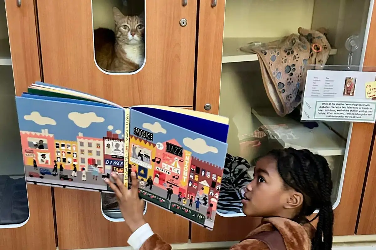 a young girl holds a book up to a cage with a cat inside