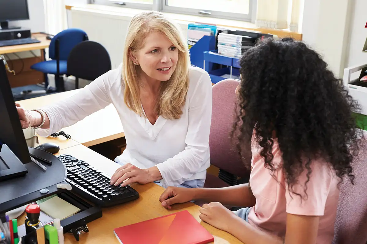a high school teacher looks at a computer screen with a student