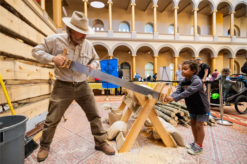 Man and small boy cutting wood with a two-person crosscut saw inside a children's museum